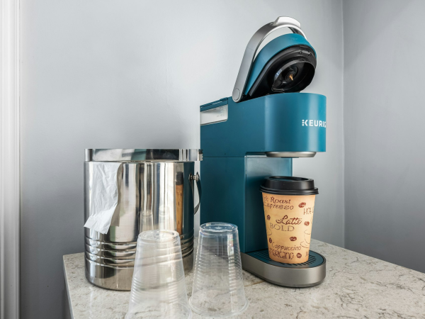 A compact coffee station with a blue Keurig, a disposable cup, glass cups, a metal waste bin, and a stack of sleeves on a marble counter.
