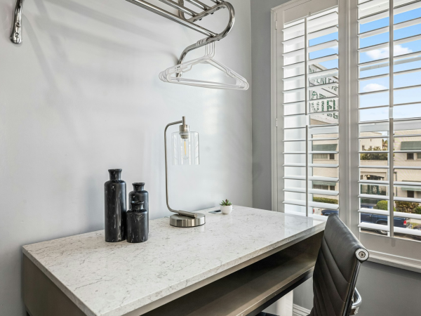 A small, modern workspace with a white speckled desk, hanging clothes rod and glassware, black decor bottles, a desk lamp, and a window with white shutters.