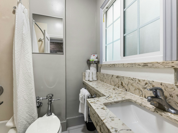 A compact bathroom with a marble countertop, double sink, beige walls, a window, a shower with a curtain, and a small shelf holding towels and toiletries.