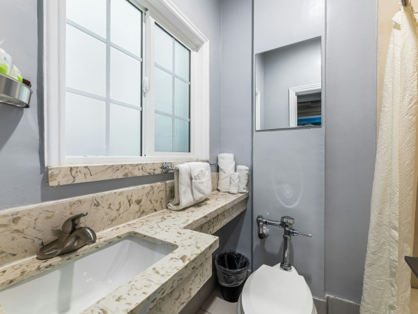 A compact bathroom with a marble vanity, double sink, stacked towels, small trash can, frosted window, light gray walls, toilet, and a shower with a beige curtain.