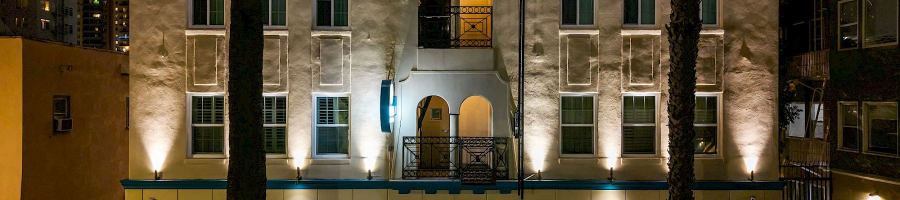 At night, a warmly lit apartment courtyard with arched entry, blue window accents, and palm trees stands between surrounding buildings.