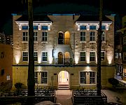 At night, a warmly lit apartment courtyard with arched entry, blue window accents, and palm trees stands between surrounding buildings.