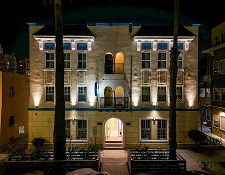 At night, a warmly lit apartment courtyard with arched entry, blue window accents, and palm trees stands between surrounding buildings.