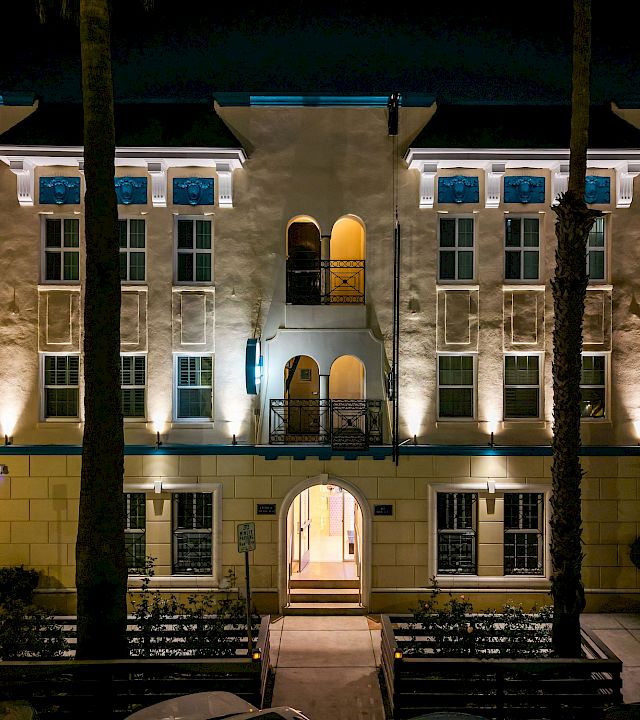 At night, a warmly lit apartment courtyard with arched entry, blue window accents, and palm trees stands between surrounding buildings.