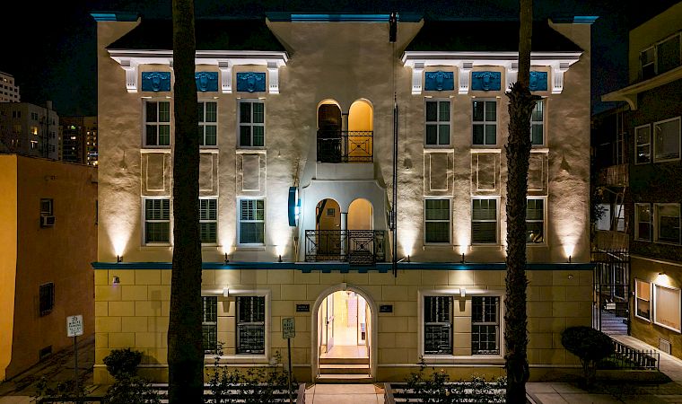 At night, a warmly lit apartment courtyard with arched entry, blue window accents, and palm trees stands between surrounding buildings.