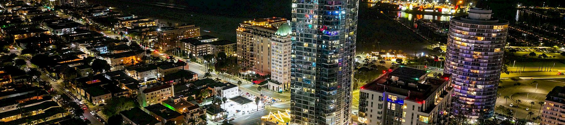 Aerial night view of a waterfront city, with illuminated high-rise hotels and apartment towers lining streets beside a dark marina.
