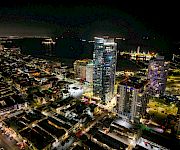 Aerial night view of a waterfront city, with illuminated high-rise hotels and apartment towers lining streets beside a dark marina.