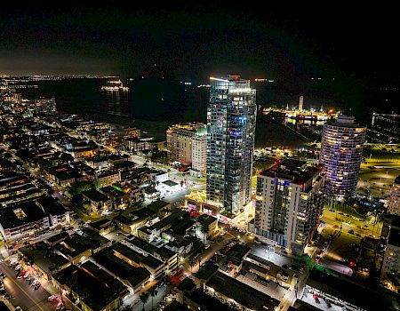 Aerial night view of a waterfront city, with illuminated high-rise hotels and apartment towers lining streets beside a dark marina.