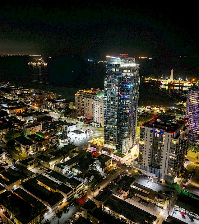 Aerial night view of a waterfront city, with illuminated high-rise hotels and apartment towers lining streets beside a dark marina.