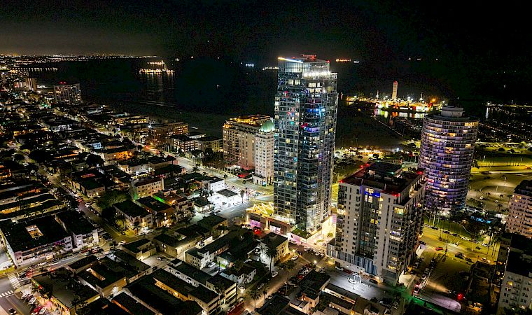 Aerial night view of a waterfront city, with illuminated high-rise hotels and apartment towers lining streets beside a dark marina.