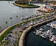 A riverside marina with yachts, a curved seawall, palm-lined roads, and high-rise skyline under a cloudy sky.